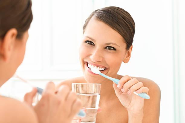 a woman brushing her teeth in front of the mirror while holding a glass of water